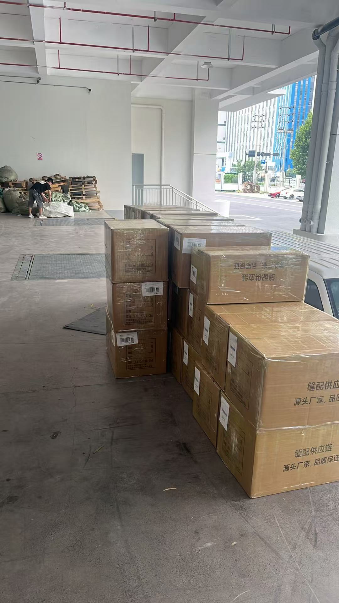 Stacks of taped cartons with barcode labels lined along a warehouse loading dock; a worker handles bagged freight and pallets in the background.