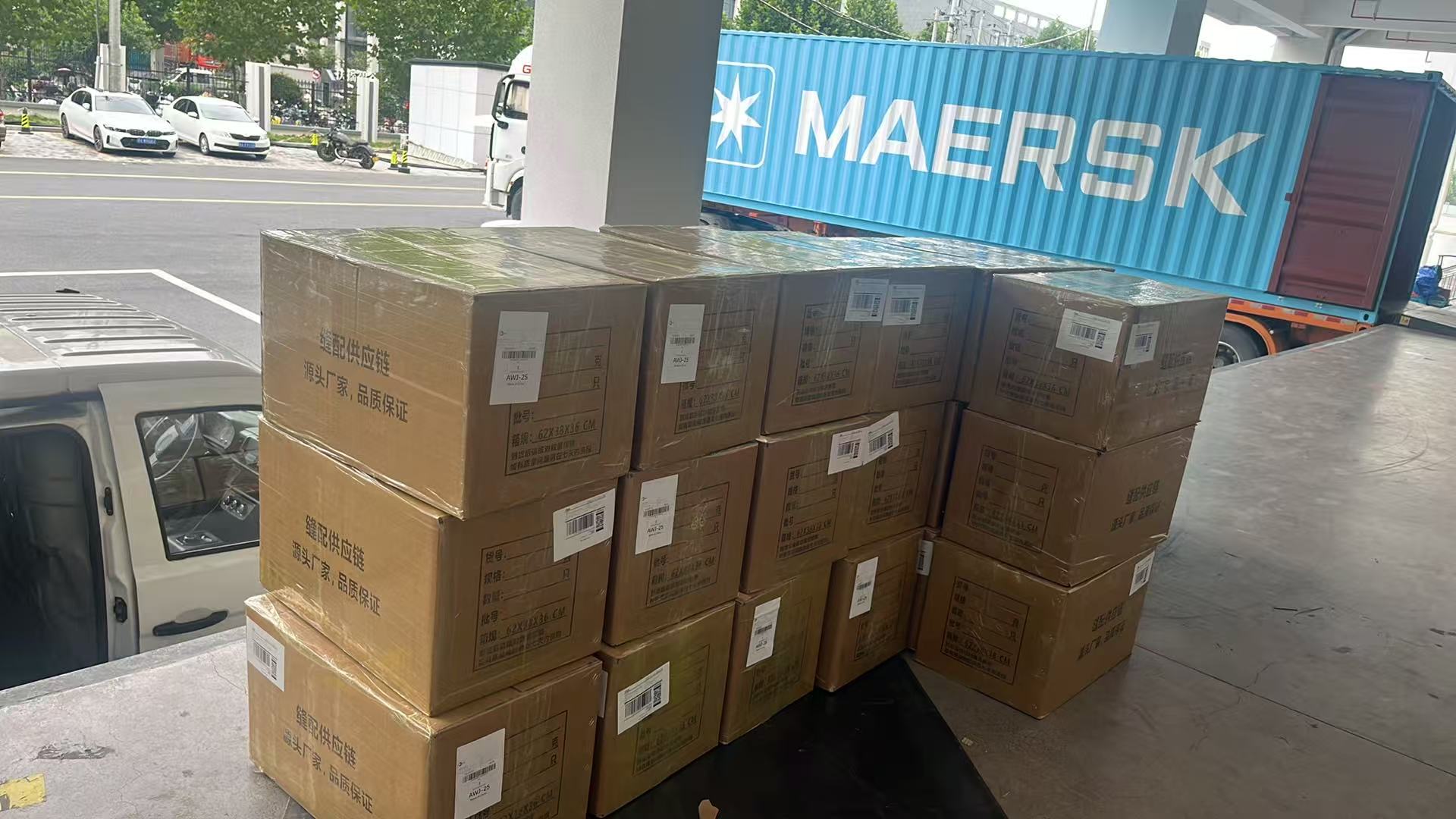 Stacked cardboard cartons with barcodes on a loading dock, with a MAERSK shipping container on a trailer in the background.