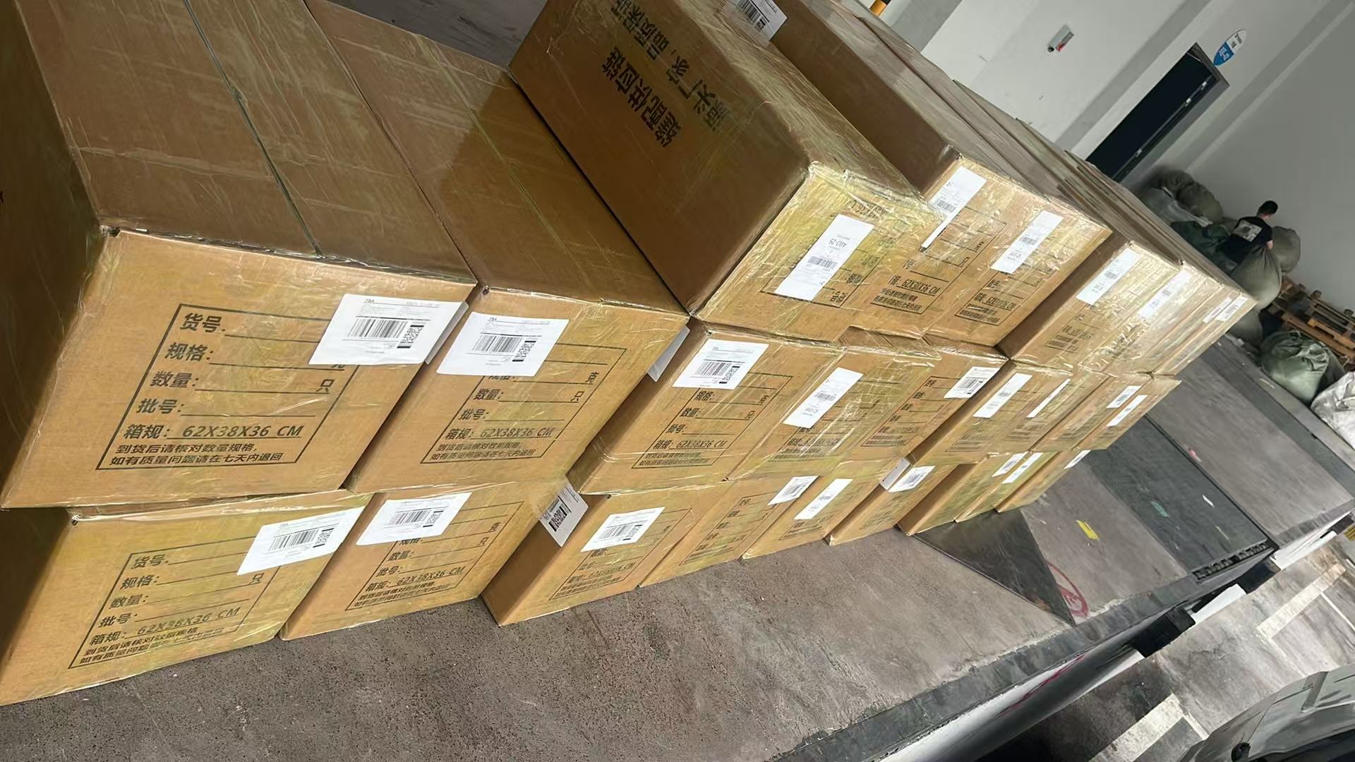 Rows of shrink-wrapped cartons with printed specs and barcode labels arranged on a dock beside a truck bay inside a warehouse.