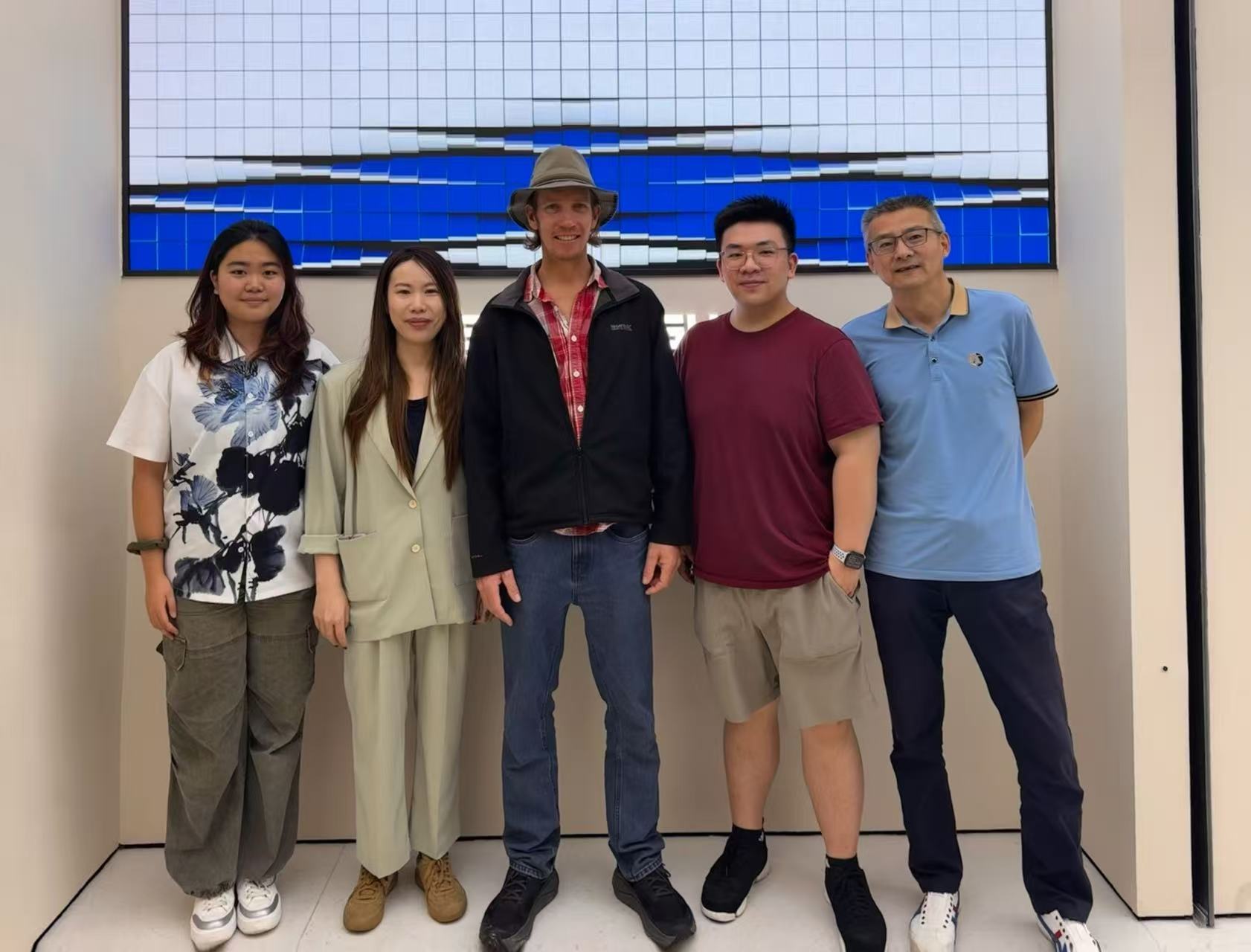 Five people stand side by side indoors, posing in front of a blue and white grid screen.