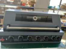 Stainless steel gas barbecue grill head with six control knobs and a lid window thermometer on a workbench in a warehouse.