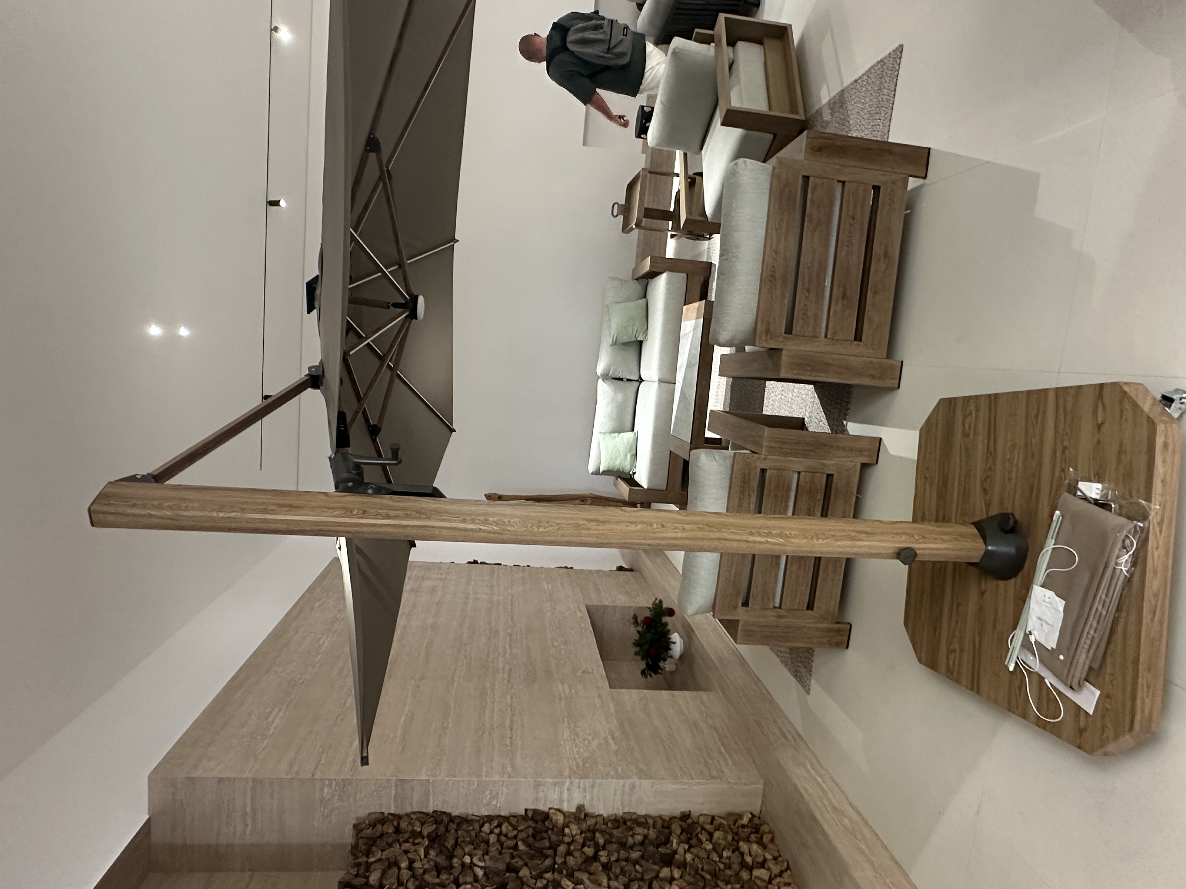 Large cantilever patio umbrella on a wheeled wooden base in a showroom, with teak-style outdoor sofas and grey-cushioned armchairs.