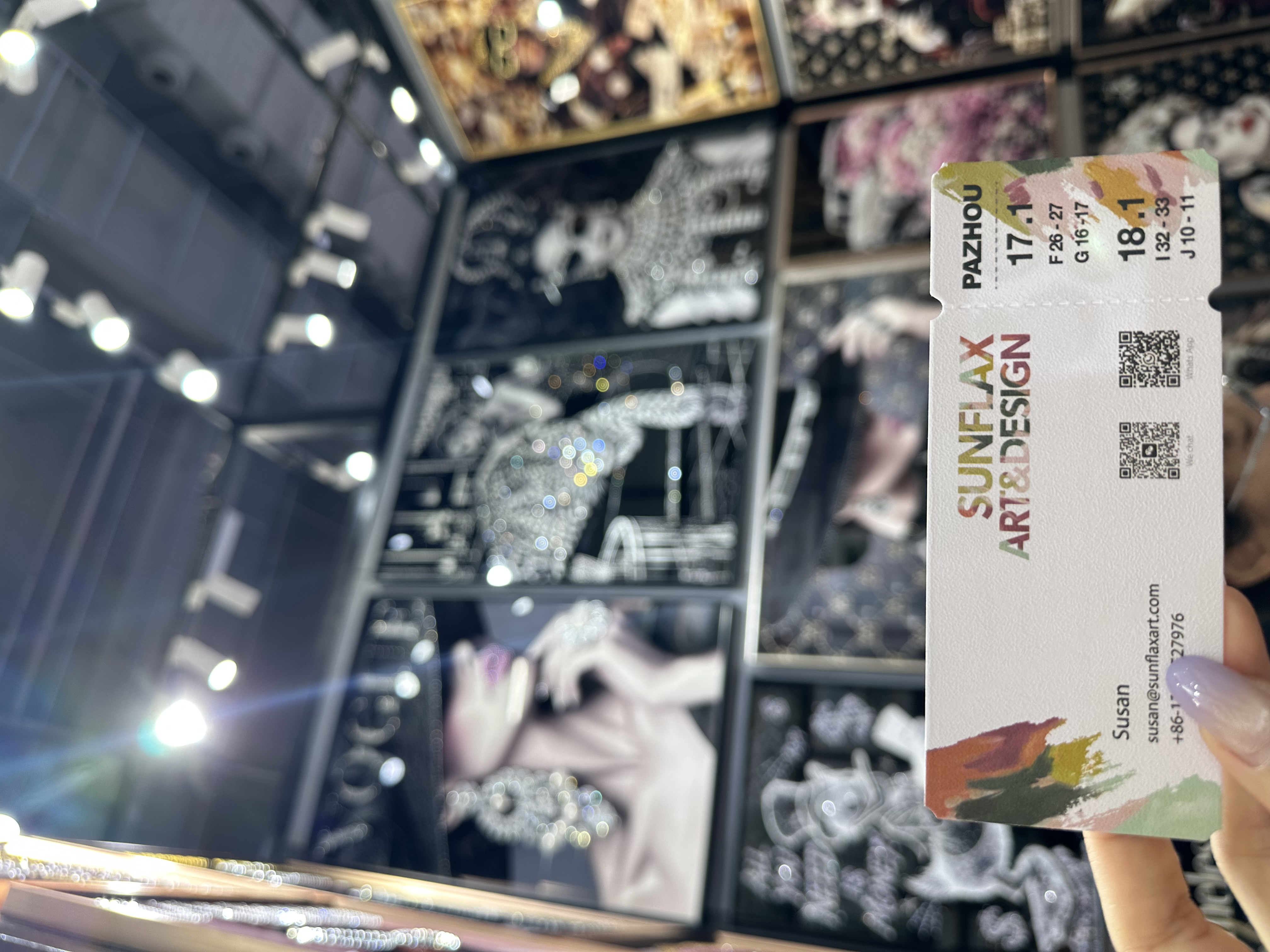 Hand holding a SUNFLAX ART & DESIGN business card at an indoor art booth with fashion-themed wall art and ceiling spotlights.