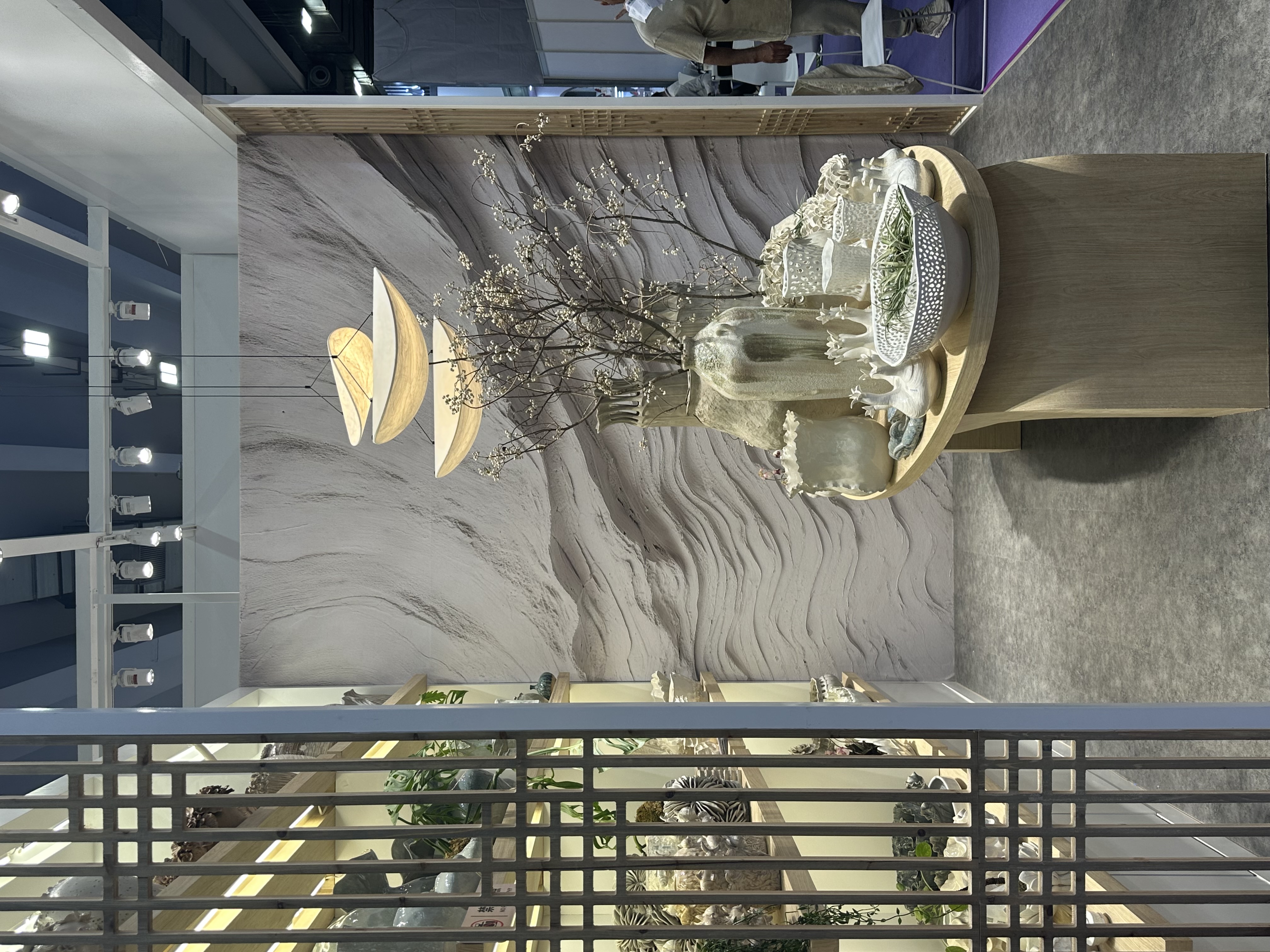 Trade fair booth displays hanging pendant lights and a table filled with white ceramic vases and bowls, with shelves of decor and a textured wall backdrop.