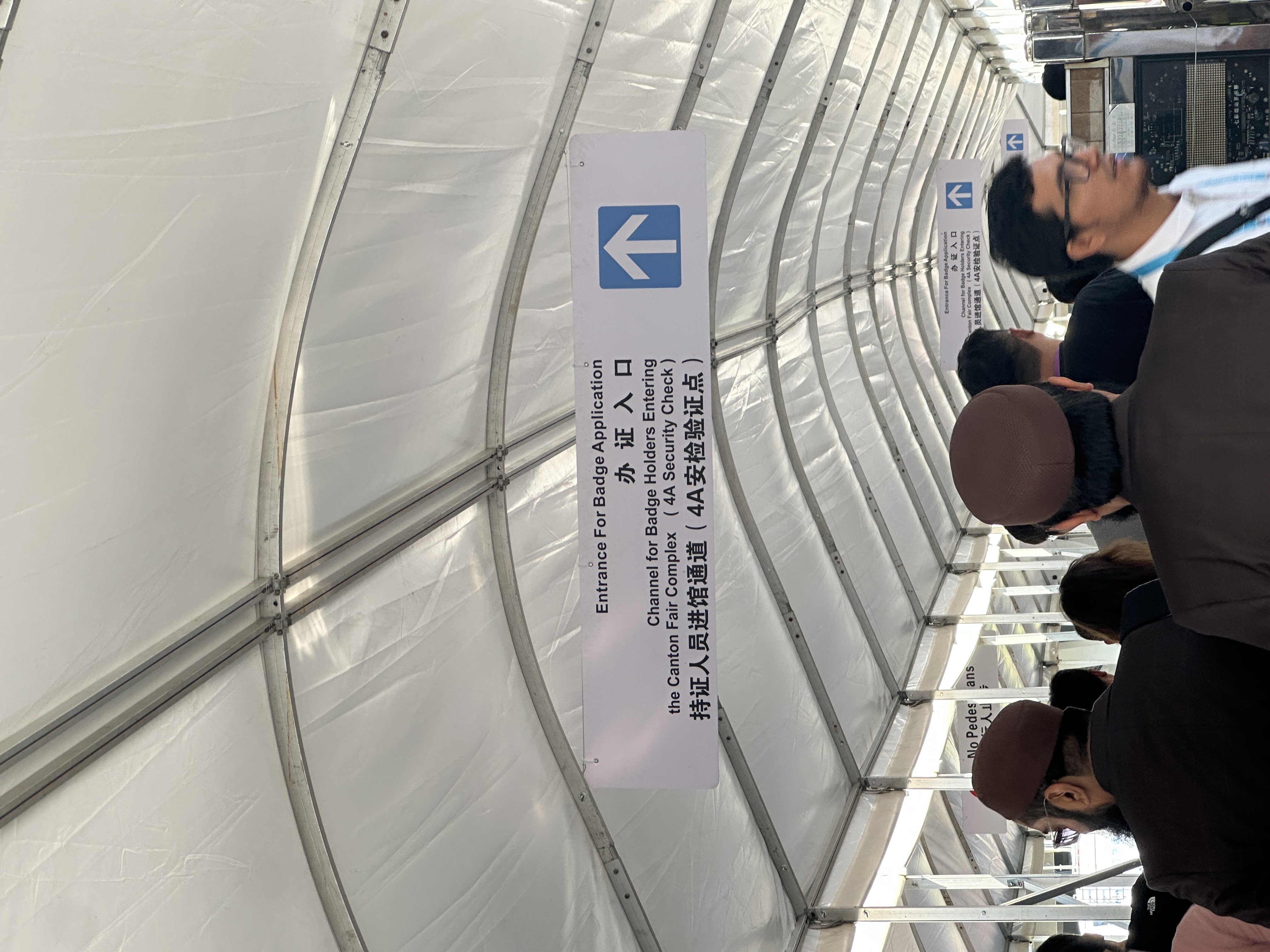 Crowd moves under a tented walkway toward signs for Entrance for Badge Application and Channel for Badge Holders entering the Canton Fair Complex.