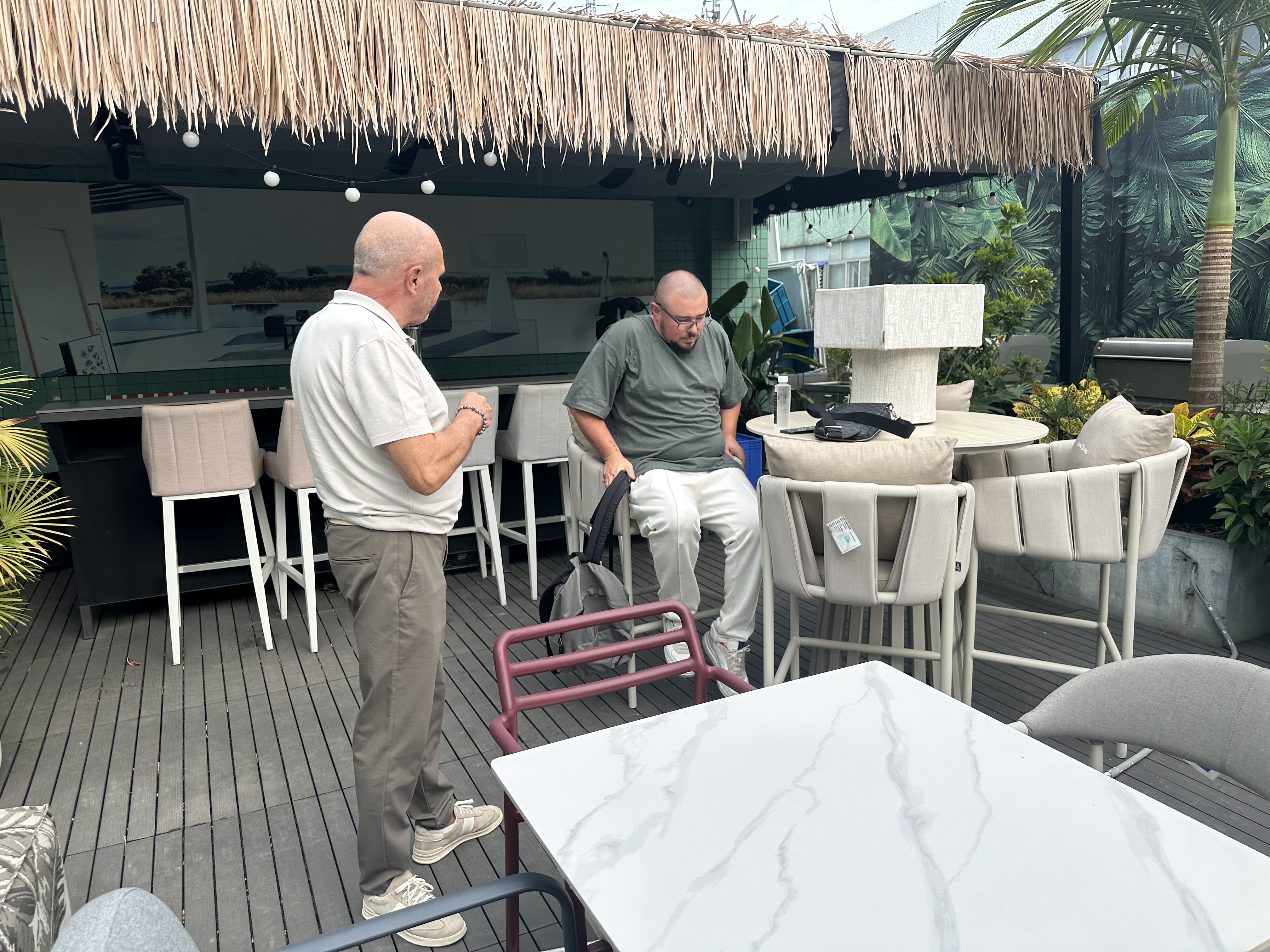 Two men in an outdoor showroom with beige bar stools, a round table on a stone pedestal, and cushioned chairs under a thatched canopy amid plants.