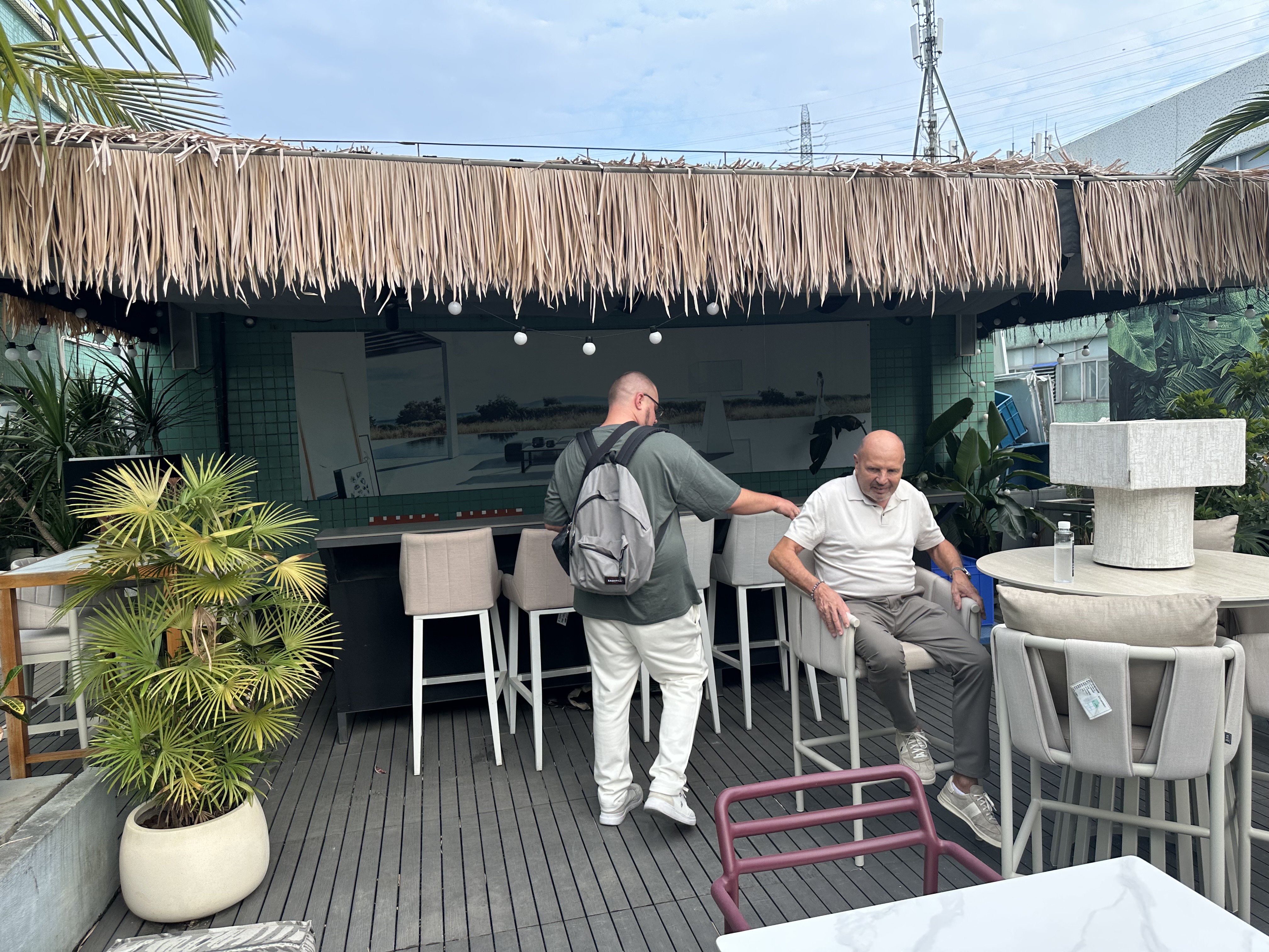 Outdoor patio furniture display with a thatched-roof bar, high stools, a round table, potted palms, and two men trying the seating.