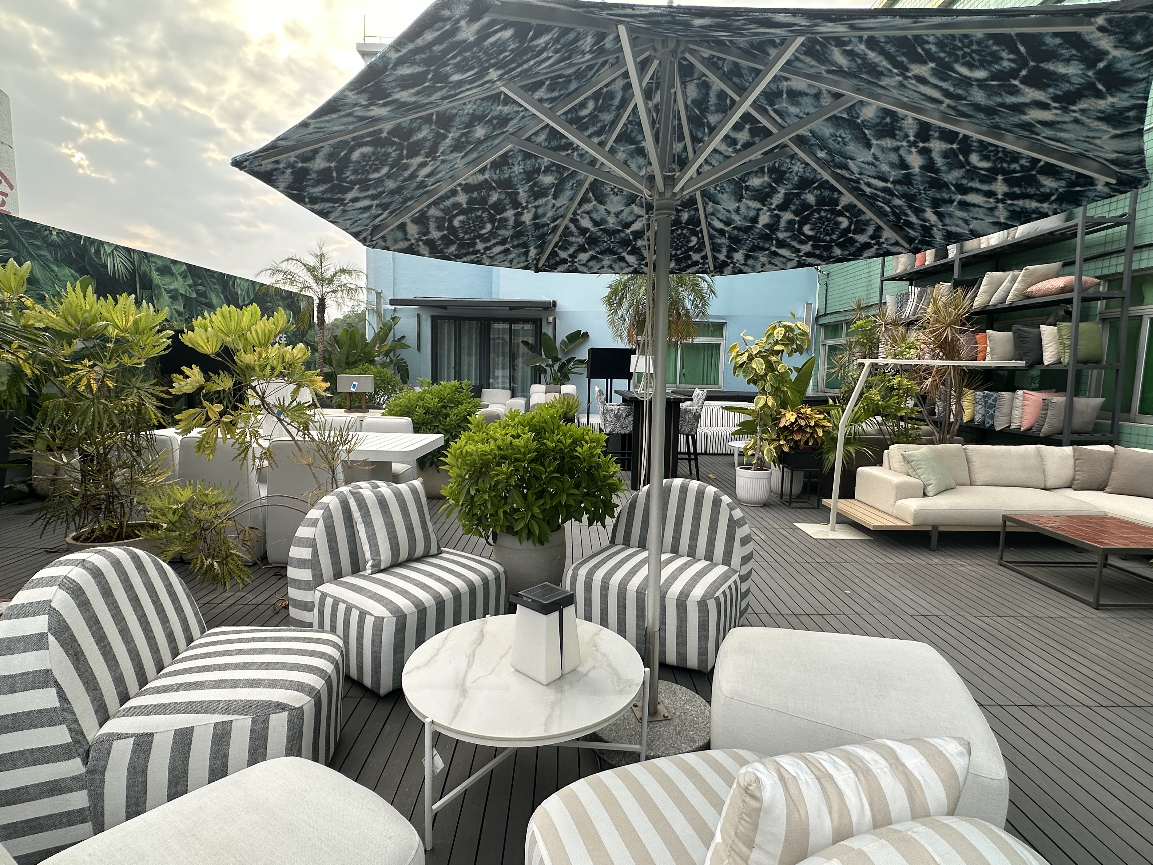 Rooftop deck display with striped lounge chairs around a small marble table, a large patterned patio umbrella, potted shrubs, sofas, and shelves of cushions.